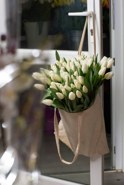 Composition with beautiful fresh flowers white tulips in eco bag hanging on door handle. Template for print. Spring shopping. - Foto, Imagem