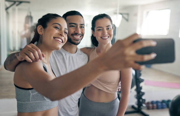 En línea para difundir un poco de inspiración fitness. Tiro de tres jóvenes atletas tomando una selfie mientras están juntos en el gimnasio. - Foto, Imagen