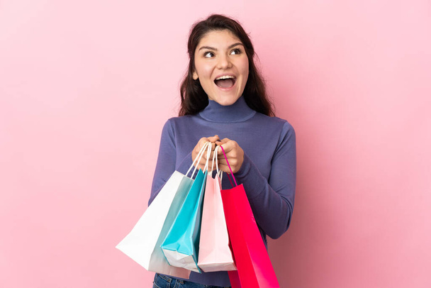 Teenager Russian girl isolated on pink background holding shopping bags and surprised - Photo, Image