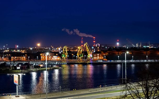 Τρεις παλαιοί λιμενικοί γερανοί φωτίζονται χρωματιστά τη νύχτα στον ποταμό Odra boulevard στο νησί Lasztownia στο Szczecin της Πολωνίας. Από απόσταση που μοιάζει με δεινόσαυρους. - Φωτογραφία, εικόνα