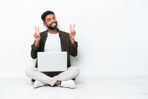 Mladý brazilský muž s laptopem, který sedí na podlaze izolovaný na bílém a ukazuje vítězné znamení oběma rukama. - Fotografie, Obrázek