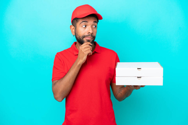 Pizza delivery Brazilian man with work uniform picking up pizza boxes isolated on blue background having doubts and thinking - Foto, immagini