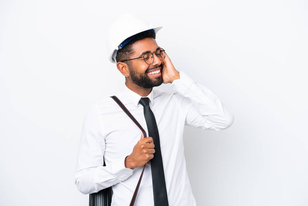 Jonge architect Braziliaanse man met helm en het houden van blauwdrukken geïsoleerd op witte achtergrond glimlachen veel - Foto, afbeelding