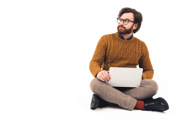 Bearded man wearing modern glasses sitting with crossed legs and holding laptop on the knees looking away from the camera - Fotó, kép