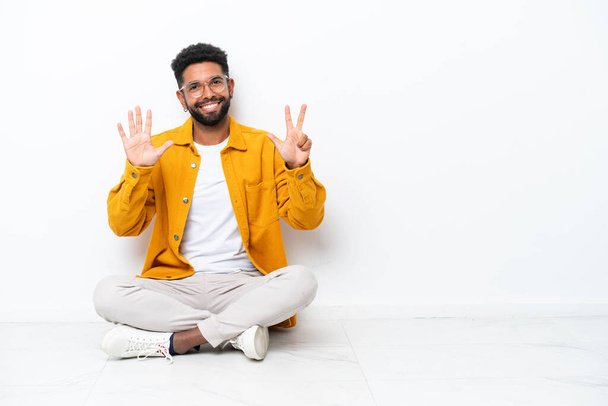 Junger brasilianischer Mann sitzt isoliert auf dem Boden auf weißem Hintergrund und zählt mit den Fingern acht - Foto, Bild