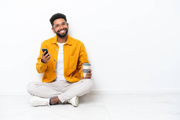 Fiatal brazil férfi ül a padlón elszigetelt fehér háttér kezében kávé elvitelre és egy mobil - Fotó, kép