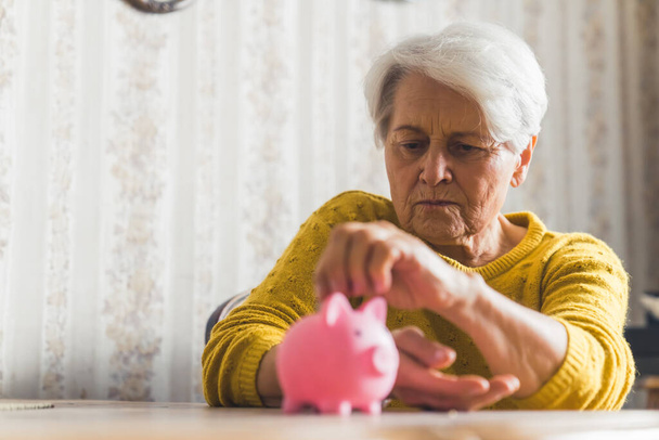 Medium indoor shot of an elderly short-haired woman in a yellow sweater putting her coins into the pink piggy bank. Savings concept. - Photo, Image