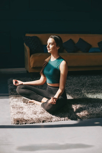 Blanke sportieve vrouw zittend in lotuspositie op een tapijt in zijn woonkamer met haar ogen dicht. Een gezonde levensstijl. Gezondheid, fitness. Meditatie - Foto, afbeelding