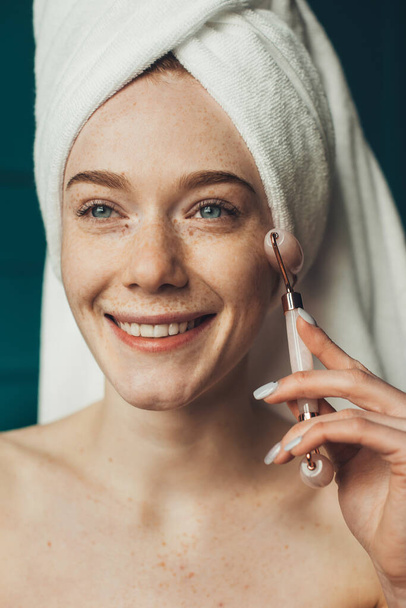Close-up portrait of smiling freckled woman holding roller massager and doing anti age facial procedure. Facial treatment. Beautiful girl. Natural stone. Skin - 写真・画像