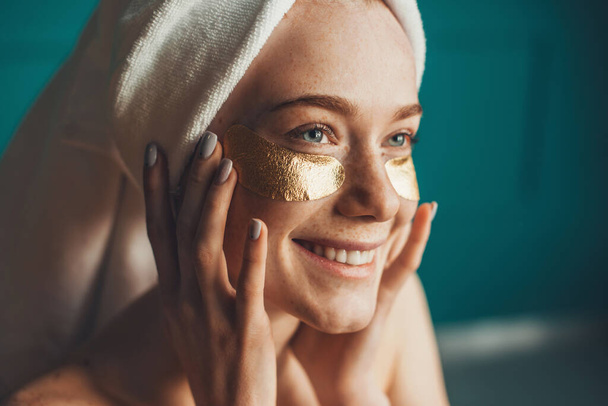 Close-up portrait of a caucasian freckled woman wearing white towel on head, with collagen golden patches under eyes. Girl facial treatment. Facial skin - 写真・画像
