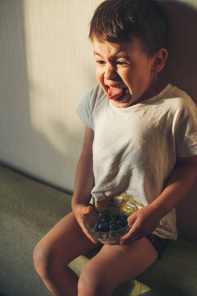 Boy eating blueberries on the couch and grimacing that he eat a sour blueberry. Facial expression. - Photo, Image