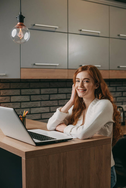 Punapäinen kaunis valkoihoinen nainen istuu kannettavan tietokoneen kanssa kotona keittiössä. Internet-viestintä, langaton yhteys teknologia. Sähköinen viestintä. - Valokuva, kuva