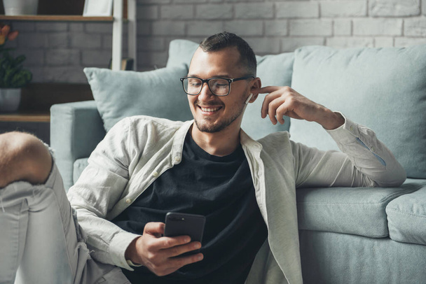 Hombre feliz usando anteojos usando el teléfono celular mientras se relaja en el suelo en casa, escuchando musicaciones en auriculares inalámbricos. Uso de auriculares y teléfono móvil - Foto, Imagen
