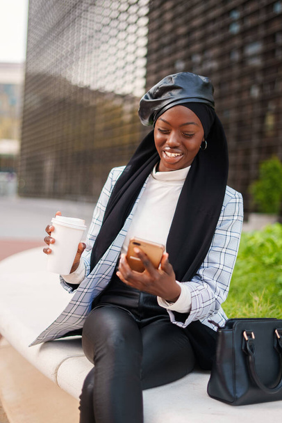 Cheerful stylish black woman in headscarf using smartphone in city - 写真・画像