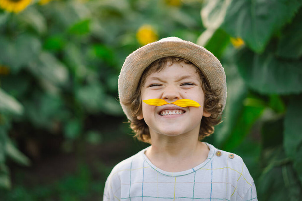 Щасливий маленький хлопчик ходить по полю соняшників і робить вуса з пелюсток соняшнику
. - Фото, зображення