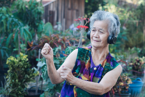 Senior Asian woman wearing wireless headphones listening to a favorite song and exercises while standing in a garden. Concept of aged people and relaxation. - Photo, Image