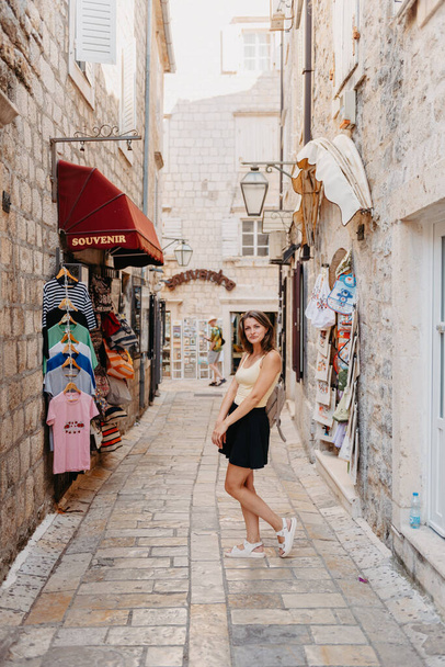 Girl Tourist Walking Through Ancient Narrow Street On A Beautiful Summer Day In MEDITERRANEAN MEDIEVAL CITY, OLD TOWN BUDVA, MONTENEGRO. Young Beautiful Cheerful Woman Walking On Old Street At - Foto, imagen