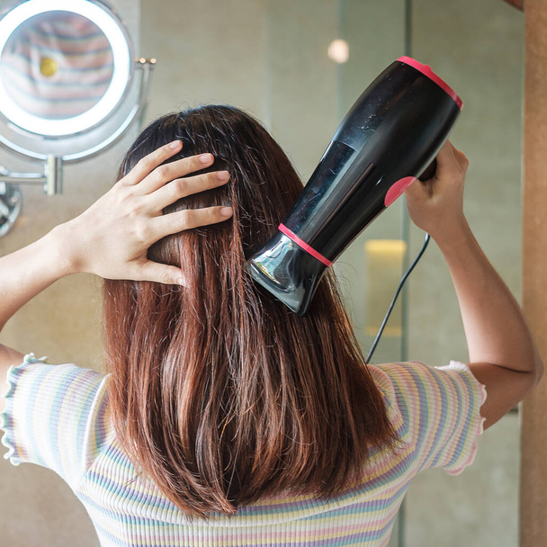 νεαρή γυναίκα που χρησιμοποιεί στεγνωτήρα μαλλιών κοντά στον καθρέφτη στο σπίτι ή το ξενοδοχείο. Χτενίσματα και έννοιες τρόπου ζωής - Φωτογραφία, εικόνα