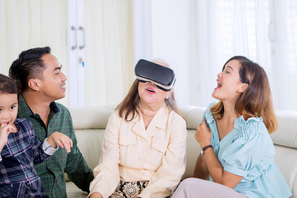 Old woman wearing VR goggles while playing with her family in the living room. Shot at home - Zdjęcie, obraz
