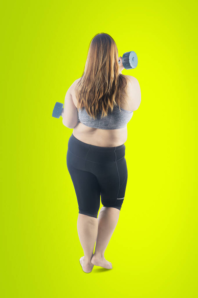 Rear view of fat woman exercising with two dumbbells while standing in the studio with green screen background - Zdjęcie, obraz