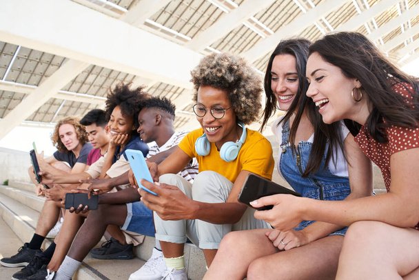 Grupa młodych mieszanych ludzi rasy z telefonami komórkowymi. Podekscytowani studenci korzystający ze swoich urządzeń technologicznych. Pojęcie młodego przedsiębiorczego, przyjaznego, selfie, app, hipster, millennial. - Zdjęcie, obraz