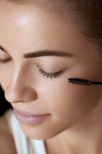 Mascara. Woman applying black mascara on eyelashes with makeup brush. Beautiful young woman face with natural eyebrows and eyelashes.   - Valokuva, kuva