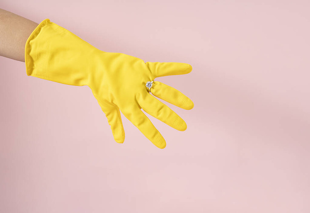 An engagement ring on the hand of a housewife. Taking care of the family is a job. Equality and women's rights. Yellow cleaning gloves on pastel pink background. - Photo, Image