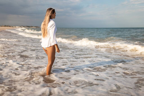 A tanned slender girl in a bluish swimsuit and a white light shirt, enjoys a warm sunny summer on the coast of a raging blue sea - Photo, image