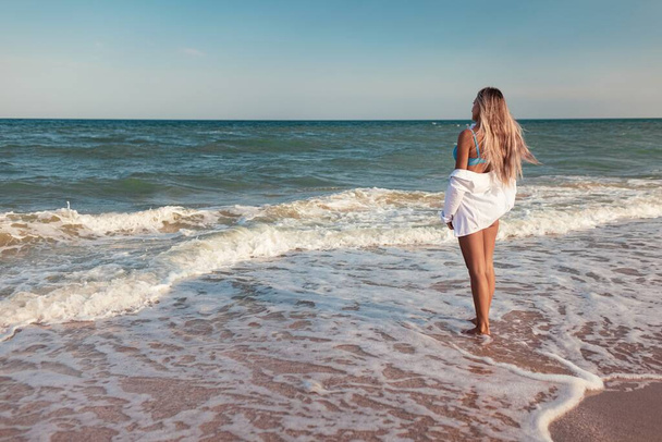 A tanned slender girl in a bluish swimsuit and a white light shirt, enjoys a warm sunny summer on the coast of a raging blue sea - Fotoğraf, Görsel