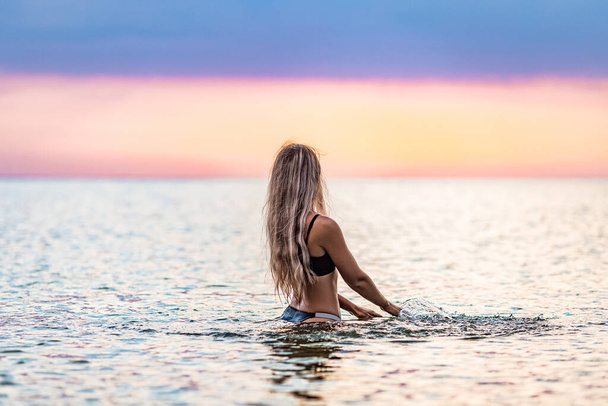 A slender tanned young girl with blond lush hair in a black swimsuit, splashes to the sides while sitting in a shallow estuary against the background of a bright warm sunset - Fotografie, Obrázek