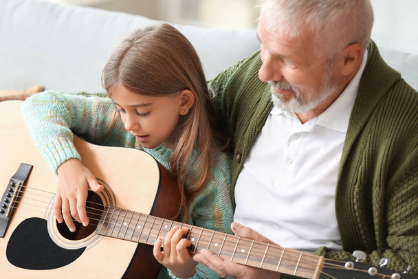 Старший чоловік навчає свою маленьку онучку грати на гітарі вдома
 - Фото, зображення