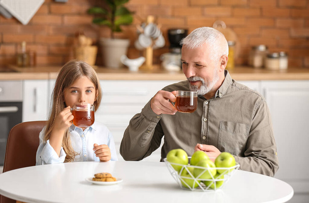 Little girl with her grandfather drinking tea in kitchen - Фото, изображение