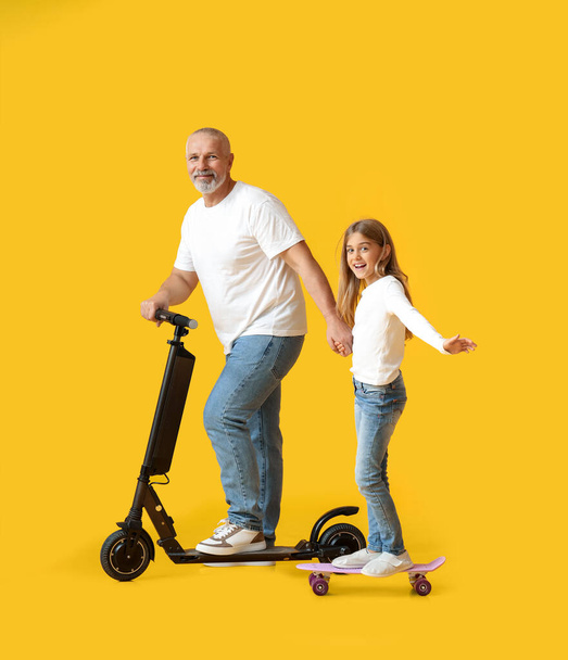 Μικρό κορίτσι και ο παππούς της με ηλεκτρικά σκούτερ και skateboard σε κίτρινο φόντο - Φωτογραφία, εικόνα