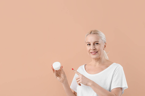 Mature blonde woman pointing at jar of cream on beige background - Photo, image