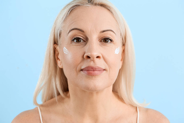 Ältere blonde Frau mit Gesichtscreme auf blauem Hintergrund, Nahaufnahme - Foto, Bild