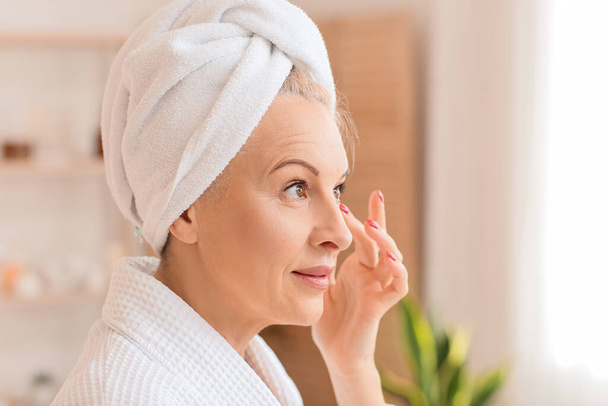 Beautiful mature woman with towel applying cream in bathroom, closeup - Foto, afbeelding