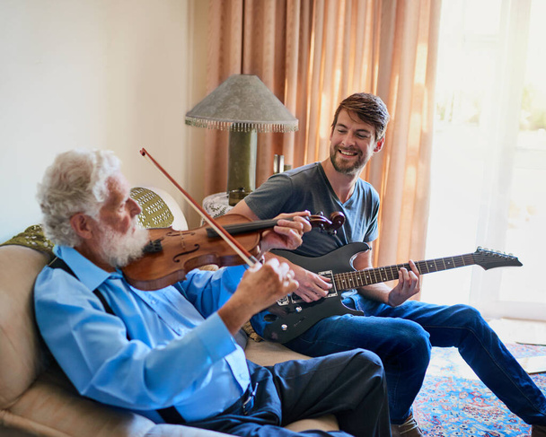 彼らの音楽を通して絆。おじいさんがソファでバイオリンを弾いている間、若い男がエレキギターを弾いている. - 写真・画像