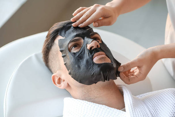 Cosmetologist applying sheet mask onto young man's face in salon, closeup - Photo, Image