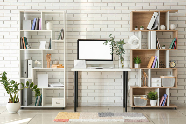 Workplace with computer, plant branches in vase and shelving units near white brick wall - Foto, Imagen