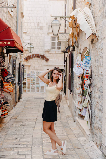 Girl Tourist Walking Through Ancient Narrow Street On A Beautiful Summer Day In MEDITERRANEAN MEDIEVAL CITY, OLD TOWN BUDVA, MONTENEGRO. Young Beautiful Cheerful Woman Walking On Old Street At - Foto, Imagen