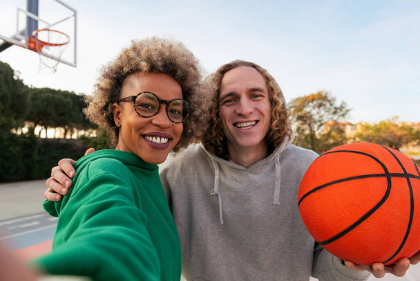 selfie of a latin woman and caucasian man smiling happy after playing in a city park basketball court, concept of friendship and urban sport in the street - 写真・画像