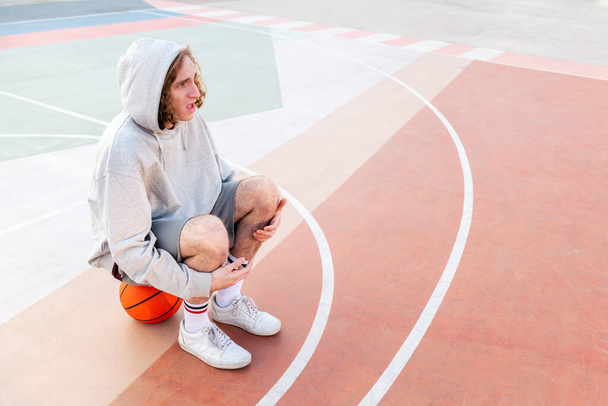 surprised caucasian man sitting on the court after basketball practice, concept of urban sport outdoors, copy space for text - Fotó, kép