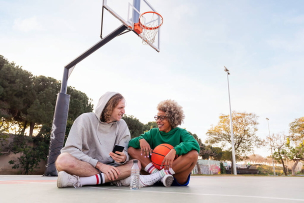 couple of friends laughing and having fun sitting on the court after a basketball practice at a city park, concept of friendship and urban sport in the street, copy space for text - Фото, изображение