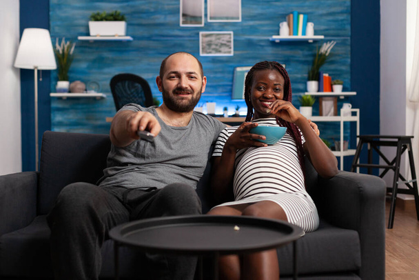 Happy interracial couple watching TV show while sharing a bowl of popcorn - 写真・画像