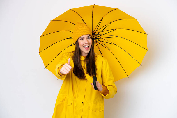 Young Brazilian woman with rainproof coat and umbrella isolated on white background with thumbs up because something good has happened - 写真・画像