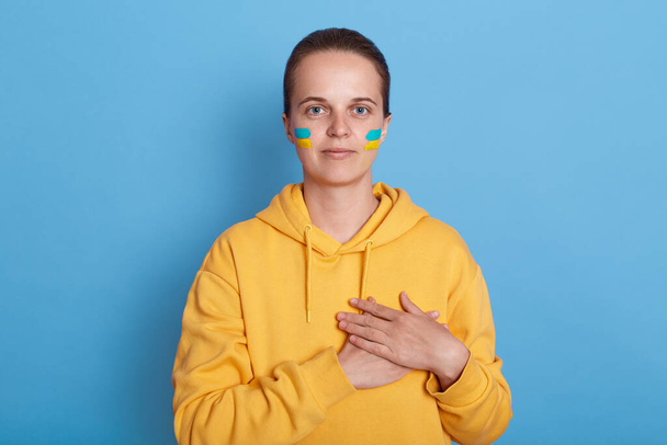 Young adult kind woman wearing yellow hoodie and with flag of Ukraine on her cheek, keeping hand on her chest, being with Ukrainians during war, isolated over blue background. Glory to Ukraine. - Fotografie, Obrázek