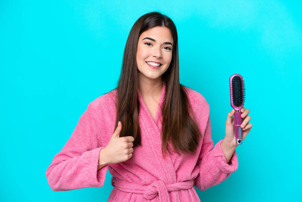 Young Brazilian woman holding hairbrush isolated on blue background with thumbs up because something good has happened - Photo, Image