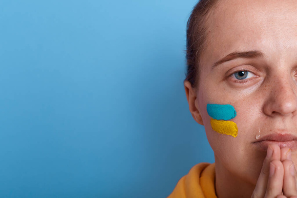 Closeup portrait of half of woman face wearing yellow hoodie and with flag of Ukraine on her cheek, female praying for Ukraine, copy space for advertisement or promotional text. Glory to Ukraine. - Φωτογραφία, εικόνα