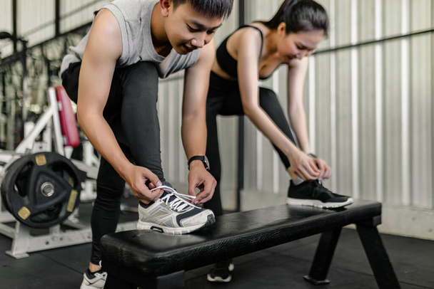 koncepce cvičení Muž a žena ve sportovním klubu spočívá jednu z jejich nohou na černé lavičce a snaží se uvázat tkaničky. - Fotografie, Obrázek
