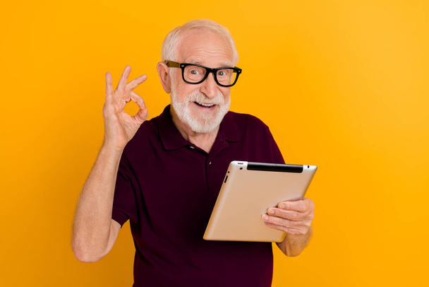 Photo of cheerful mature man show fingers okey symbol approve good tablet app isolated over yellow color background - Foto, Imagem